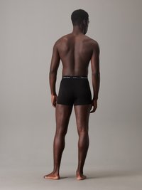 Schwarze Boxershorts aus glattem Stoff, mit einem elastischen Bund mit Logo, getragen von einer Person, die von hinten steht.