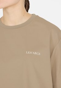 Beige puuvillane t-särk, ümar kaelus, mille vasakul rinnal on valge "LES'ARCS" tekst. Sile tekstuur, lühikesed varrukad.