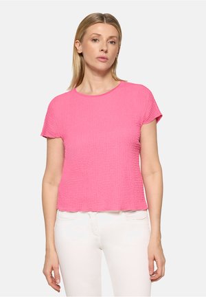 Frau trägt ein leuchtend pinkes, strukturiertes Kurzarmoberteil und weiße Hose, steht vor einem schlichten hellgrauen Hintergrund.