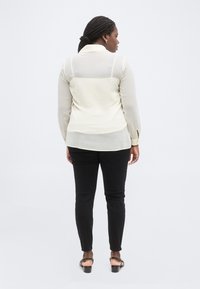 Γυναίκα με πλεγμένα μαλλιά που φοράει διάφανη κρεμ μπλούζα, μαύρο παντελόνι και μαύρα σανδάλια, στέκεται με την πλάτη προς το λευκό φόντο.