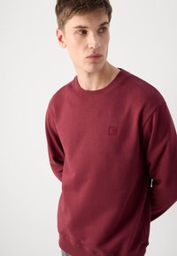 Sudadera burdeos hecha de mezcla de algodón, presenta mangas largas, un cuello redondo y un pequeño logo cuadrado bordado en el pecho. Textura suave.