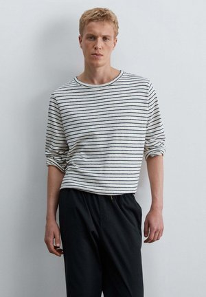 Jonge man met kort blond haar die een wit shirt met lange mouwen draagt met dunne zwarte strepen en een zwarte broek, staand tegen een eenvoudige witte muur.