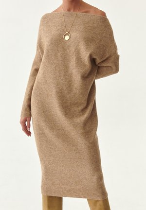 Długa beżowa sukienka typu off-shoulder, zestawiona ze złotym naszyjnikiem z okrągłą zawieszką oraz beżowymi kozakami za kolano.