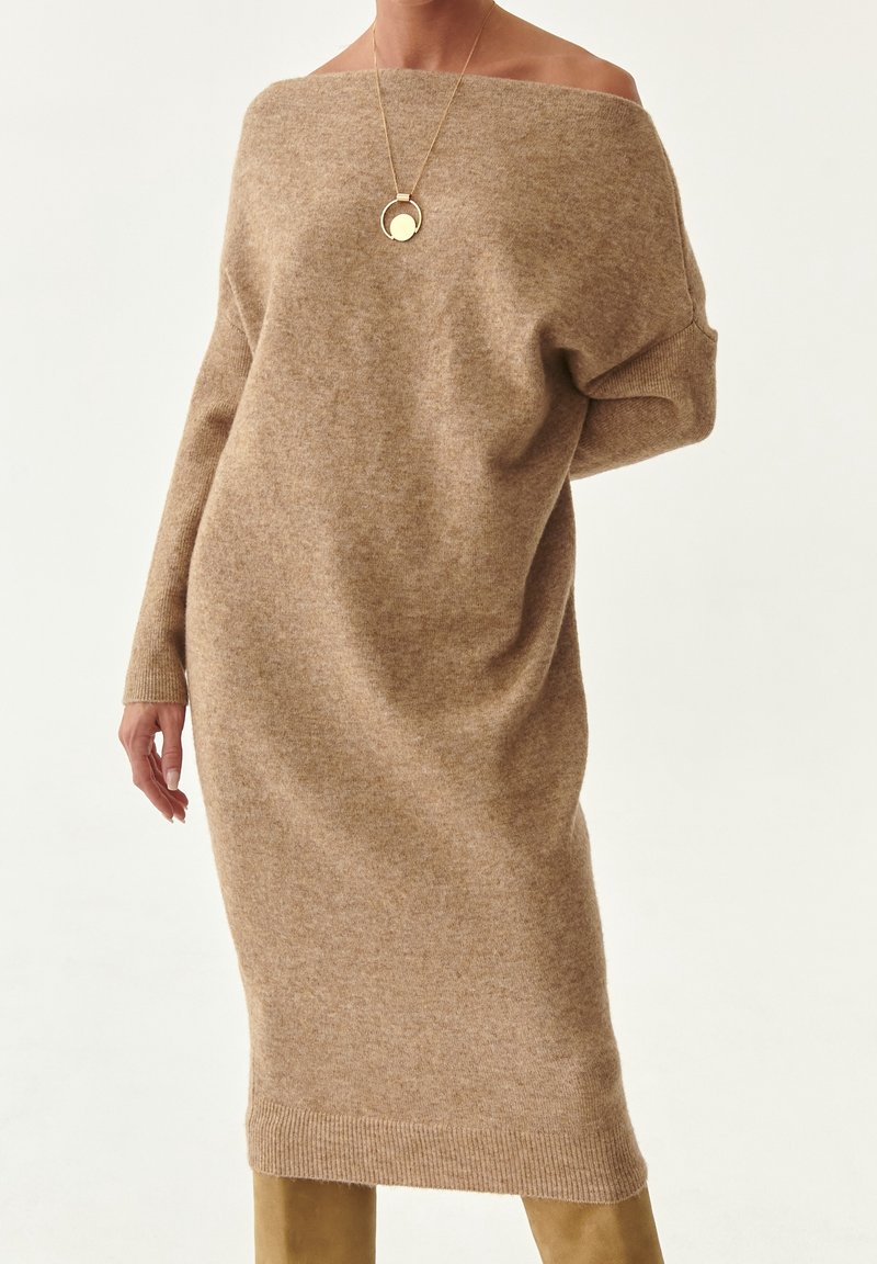 Robe pull longue beige à épaules dénudées associée à un collier pendentif circulaire doré et des bottes hautes jusqu'aux genoux couleur beige clair.