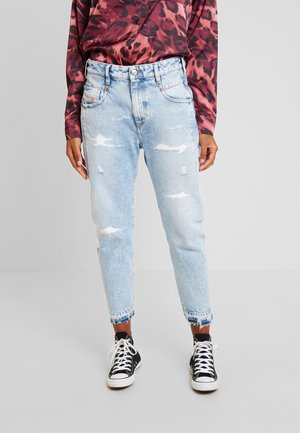 Osoba ubrana w jasnoniebieskie podarte dżinsy z postrzępionymi brzegami, czarne wysokie trampki oraz czerwoną, wzorzystą koszulę z długim rękawem.