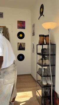 Kot prostorije s vinilnimi ploščami in albumi na steni, visoka omara za čevlje s čevlji, talna svetilka in oseba v svetlih kratkih hlačah ter beli majici.