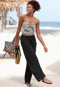 Halterloses Oberteil mit schwarz-weißen Muster und Ausschnitt-Detail, kombiniert mit locker sitzenden schwarzen Hosen und Sandalen. Trägt eine gewebte Tasche.