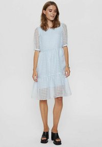 Svijetloplava haljina s kratkim, teksturiranim, napuhanim rukavima, slojevitim rubom i uzorkom houndstooth; nosi se s crnim platformama.