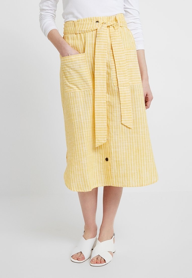 Femme portant une jupe midi rayée jaune avec des poches avant, une ceinture nouée à la taille, des sandales blanches et un haut blanc à manches longues.
