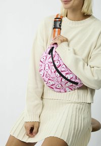 Donna che indossa un maglione lavorato a maglia color crema e una gonna, mentre tiene una borsa a tracolla rosa con facce di cani dei cartoni animati e una tracolla a righe arancioni.