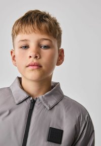 Siva lahka jakna s ovratnikom in črnim zadrgo. Ima kvadratno oznako na prsih. Gladka, sijoča tekstura tkanine.