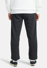 Zwarte sweatpants met een ontspannen pasvorm, elastische tailleband en een achterzak, gecombineerd met groene sneakers en witte sokken.