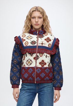 Un mannequin se tient devant un fond uni, vêtu d’une veste matelassée colorée à motifs floraux et d’un jean bleu.
