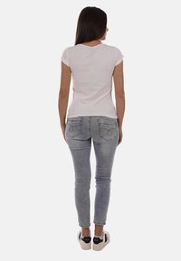Femme aux longs cheveux bruns portant un t-shirt blanc ajusté, un jean skinny gris et des baskets blanches, vue de dos.