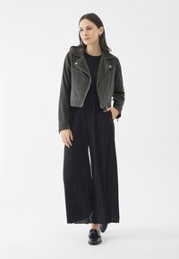 Chaqueta corta de cuero negro con cremallera plateada, combinada con pantalones anchos plisados negros y mocasines negros. Textura suave y diseño estructurado.