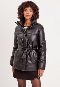 Czarna kurtka puchowa z imitacji skóry, z zatrzaskami, oversize'owy kołnierz, pasek w talii oraz pikowane wykończenie. Modelka ma na sobie czarną spódnicę.