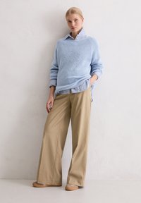 Helles, blaues Strickpullover über einem gestreiften Hemd, kombiniert mit lockeren beige Hosen und braunen Schuhen, vor einem neutralen Hintergrund.