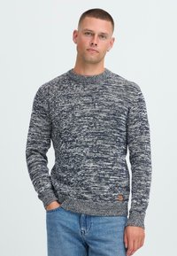 Jaunas vyras su trumpais plaukais, vilkintis tamsiai mėlyną ir baltą įstrižų raštų megztinį ir mėlynas džinsus, stovi su viena ranka kišenėje.