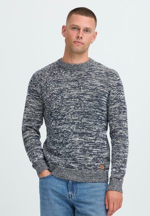 Junger Mann mit kurzgeschnittenem Haar, der einen navy-blauen und weißen melierten Strickpullover und blaue Jeans trägt und mit einer Hand in der Tasche steht.