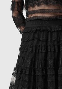 Femme portant un haut en dentelle noire transparente à manches longues et une jupe en dentelle noire à volants.
