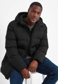 Black puffer jacket with a hood, featuring a zip front, quilted texture, and long sleeves. Worn over a gray sweater and blue jeans.