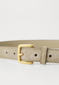 Beige nahka-vyö kultaisella metallisoljella, jossa on useita reikiä säätämistä varten ja huomaamatonta ompelua reunoilla.