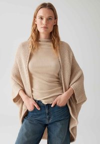 Cardigan en tricot beige avec des manches larges, porté par-dessus un haut beige à côtes et un jean bleu. Présente un tissu texturé et une coupe décontractée.