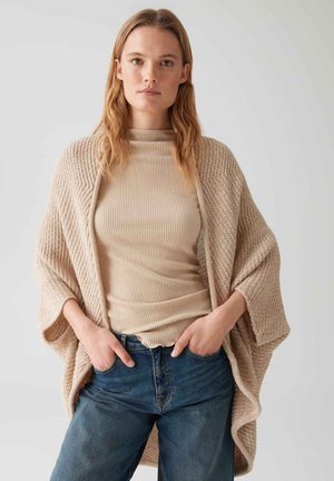Beige knitted cardigan with wide sleeves, worn over a ribbed beige top and blue jeans. Features textured fabric and a relaxed fit.