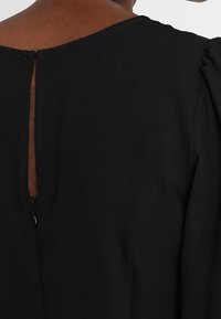 Schwarzes Kleidungsstück mit rundem Halsausschnitt und einem subtilen Schlüsselloch-Ausschnitt hinten. Mit kurzen Puffärmeln und glatter Stofftextur.