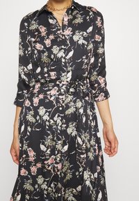 Černé šaty s květinovým vzorem, obsahujícím ptáky a zeleň. Dlouhé rukávy, zapínání na knoflíky v přední části a vázání v pase. Hedvábná textura.