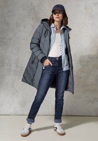 Puffy gray parka with a hood, layered over a light blue striped shirt and white tank top, paired with dark jeans and white sneakers.