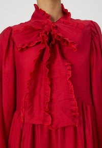 Piros ruha fodros nyakkivágással és masnival, könnyű anyagból készült. Hosszú ujjakkal és plisszírozott szoknyával rendelkezik. Strukturált felület.