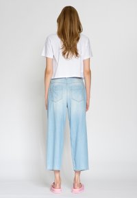 Ljusblå vida jeans med en urblekad textur, i kombination med en vit kort t-shirt och rosa slip-on sandaler. Bakifrån.