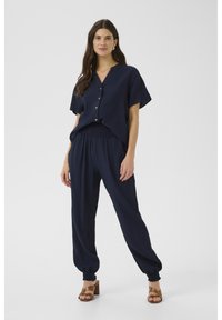 Námornícky modrý dvojdielny outfit pozostávajúci z voľného topu s krátkym rukávom a zapínaním na gombíky vpredu, a širokých nohavíc s elastickým pásom a zúženými manžetami.