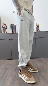 Cilvēks, kurš ģērbies gaiši pelēkās sporta biksēs un brūnās Adidas kedās, stāv uz koka grīdas blakus pelēkai skapī, uz kura atrodas krāsaini dzīvnieku skulptūras.