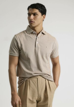 Camisa polo color beige claro con cuello, tapeta de tres botones y logotipo bordado. Llevada con pantalones plegados color beige claro. Textura lisa.