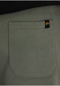 Tissu en coton vert olive avec une poche arrière cousue, ornée d'une petite étiquette noire avec un logo jaune. Texture lisse, coupe ajustée.