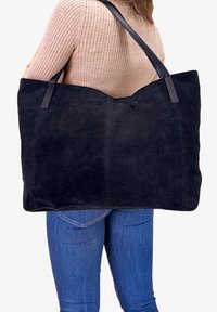 Kobieta w niebieskich dżinsach i różowym swetrze w prążki trzyma dużą czarną torebkę z zamszu na ramieniu na tle jednego, białego koloru.