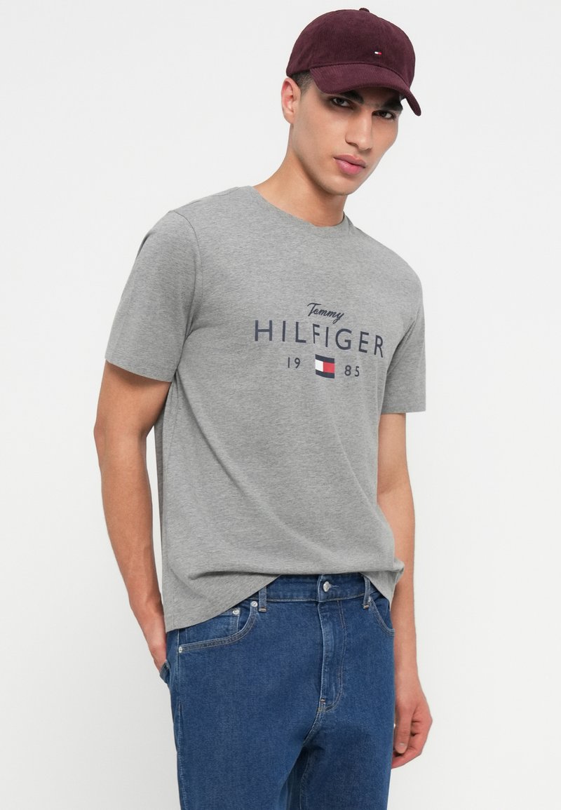Ung mand iført en grå Tommy Hilfiger t-shirt og en vinrød kasket med hænderne i lommerne på sine blå jeans mod en hvid baggrund.