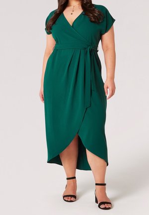 Mujer que lleva un vestido cruzado verde con mangas cortas y cintura atada, combinado con tacones negros con pulsera en el tobillo.