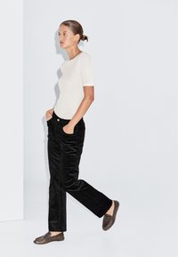 Camicetta beige a maniche corte, pantaloni a gamba larga in velluto a coste neri e mocassini marroni. Modella che cammina con le mani in tasca, sfondo minimalista.