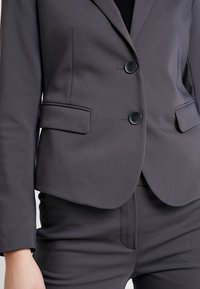 Blazer ajusté gris foncé avec une texture subtile, deux boutons avant et des poches latérales. Coupe slim avec revers à crans.