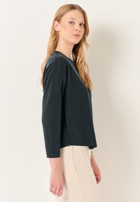 Maglietta lunga verde scuro con una vestibilità rilassata, scollatura rotonda e texture morbida. Indossata con un capo inferiore di colore chiaro.