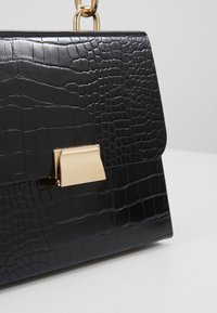 Sac à main noir à motif crocodile avec un fermoir rectangulaire doré et une anse chaîne dorée sur un fond uni clair.