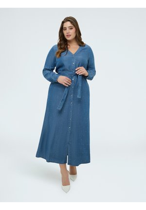 Femme portant une longue robe chemise bleue avec une ceinture à la taille et des talons blancs, debout devant un fond clair uni.