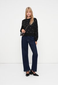 Sweatshirt noir avec motif de cœur doré, associé à un jean large bleu foncé et des chaussures à talons noires. La tenue est accessorisée d'un petit sac.