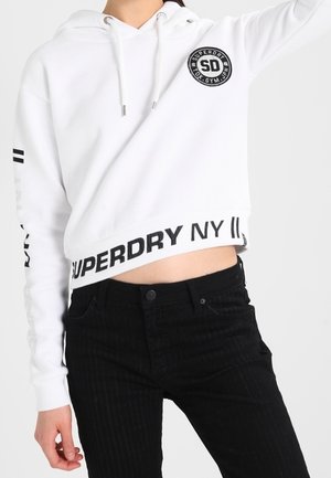 Osoba ubrana w białą krótką bluzę z kapturem z czarnym napisem "SUPERDRY NY" i czarne spodnie, podnosząca jedną rękę na jednolitym tle.