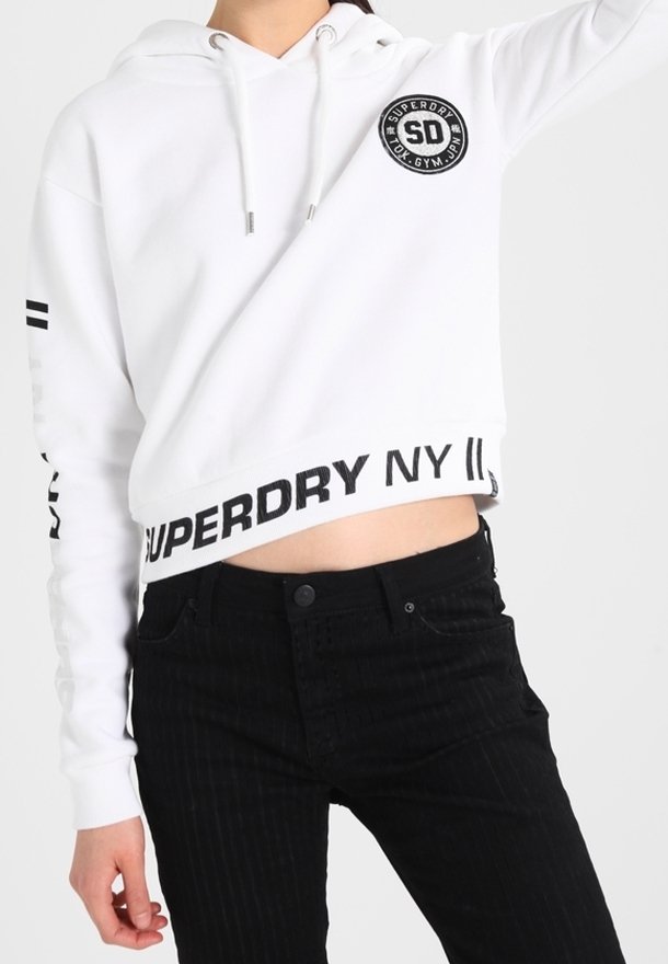 Personne portant un sweat à capuche court blanc avec le texte noir "SUPERDRY NY" et un pantalon noir, levant un bras devant un fond uni.