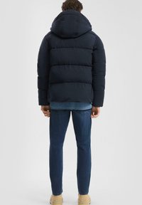 Mornarski pufasti jakna s kapuco, z značilnim prešitjem in rahlo sijočo teksturo. Nosena čez svetlo modro teksturirano plast in temne kavbojke.