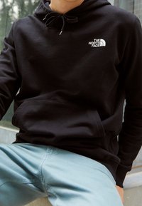 Personne portant un sweat à capuche noir The North Face avec cordons et poche avant, associé à un pantalon bleu clair, assise en extérieur.